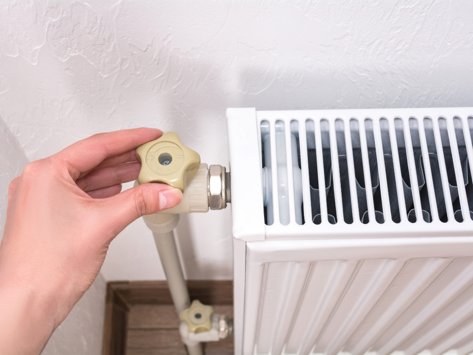 A hand adjusts the knob on a white radiator against a textured wall. The setting suggests a cozy, indoor space focused on controlling room temperature.