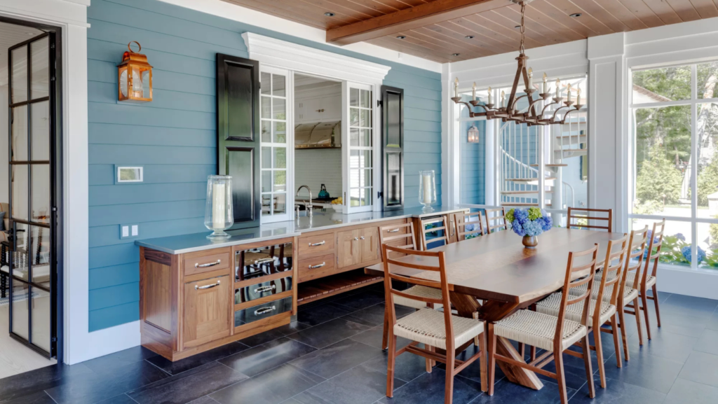 How Chris Rapczynski Retrofits Historic Buildings for Climate Resilience Spacious kitchen with blue walls, wooden cabinets, and a large dining table. A window offers garden views, and a rustic chandelier adds charm.