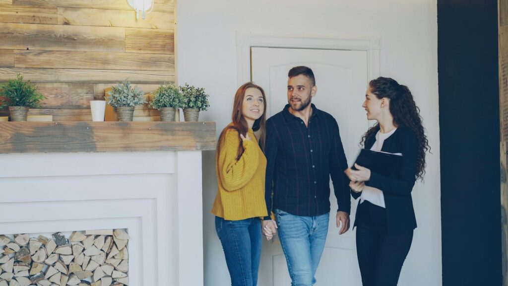 A couple discusses home options with a real estate agent in front of a cozy fireplace.